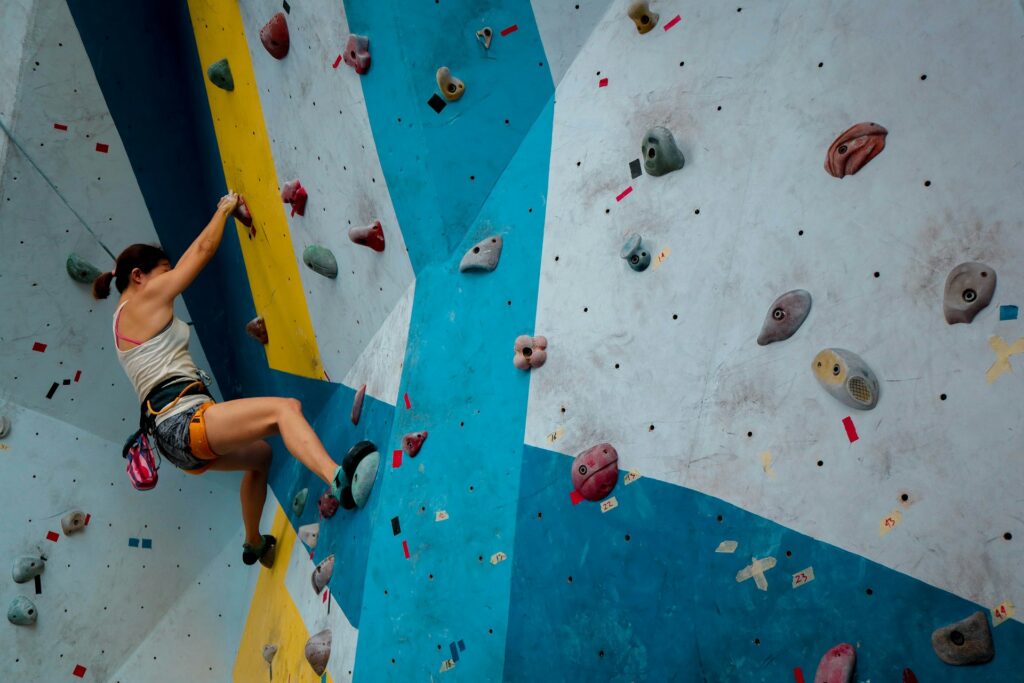 A rock climber ascends a colorful indoor wall, gripping holds of various shapes and sizes, surrounded by blue and yellow sections.