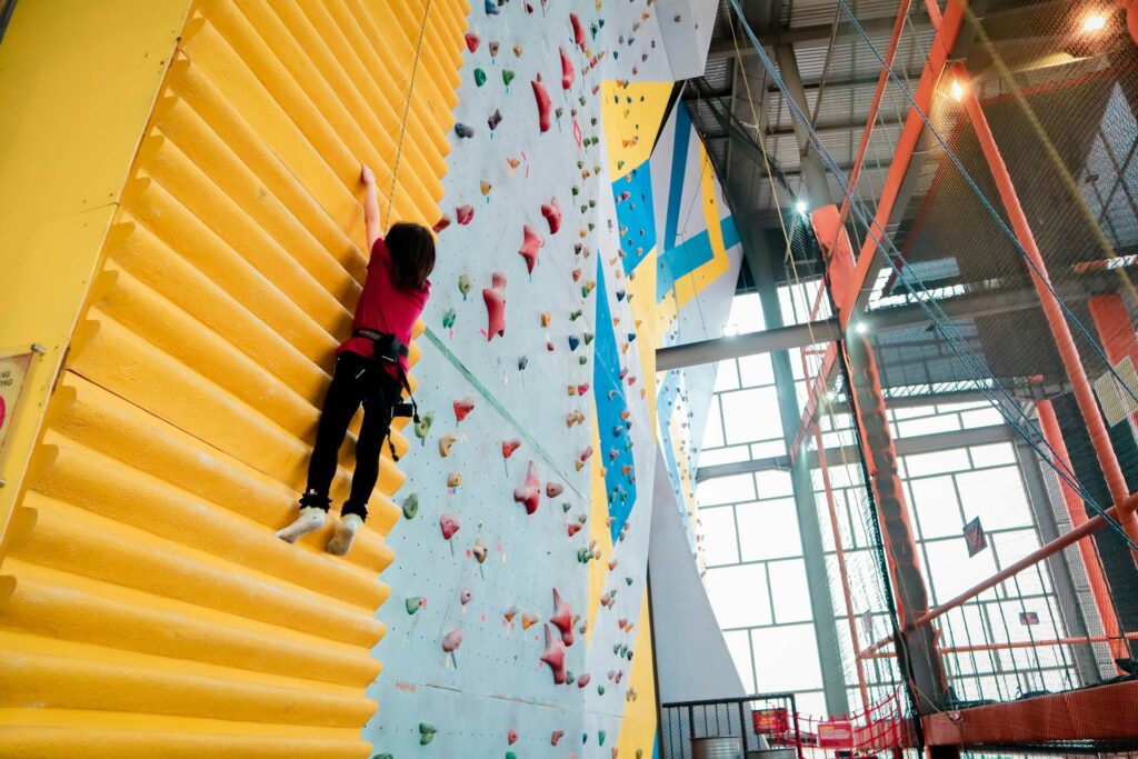 A climber in a red shirt ascends a textured yellow wall in an indoor climbing gym, with colorful rock holds on a nearby wall.