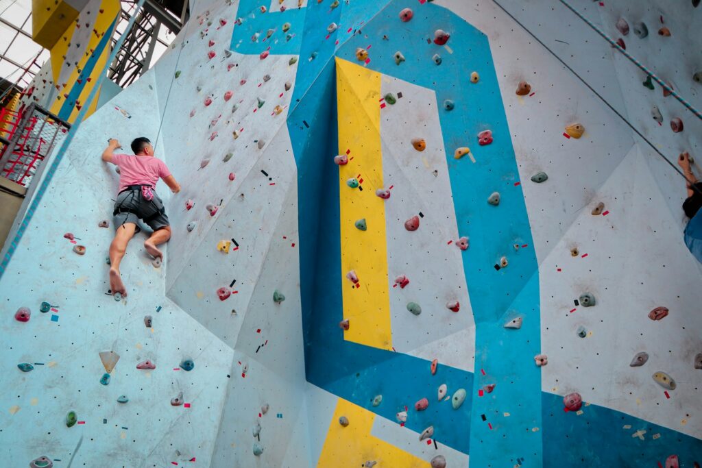 A climber in a pink shirt ascends a colorful indoor rock wall with various holds, showcasing a challenging route.