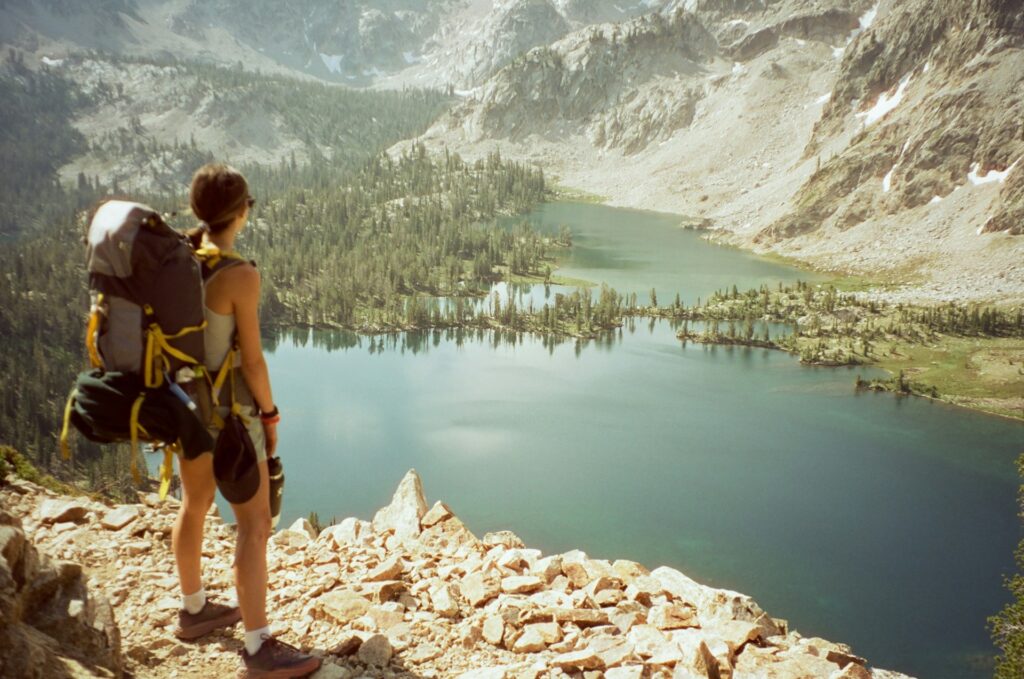 A hiker stands on rocky terrain, gazing at a serene lake surrounded by trees and mountains under a clear sky.
