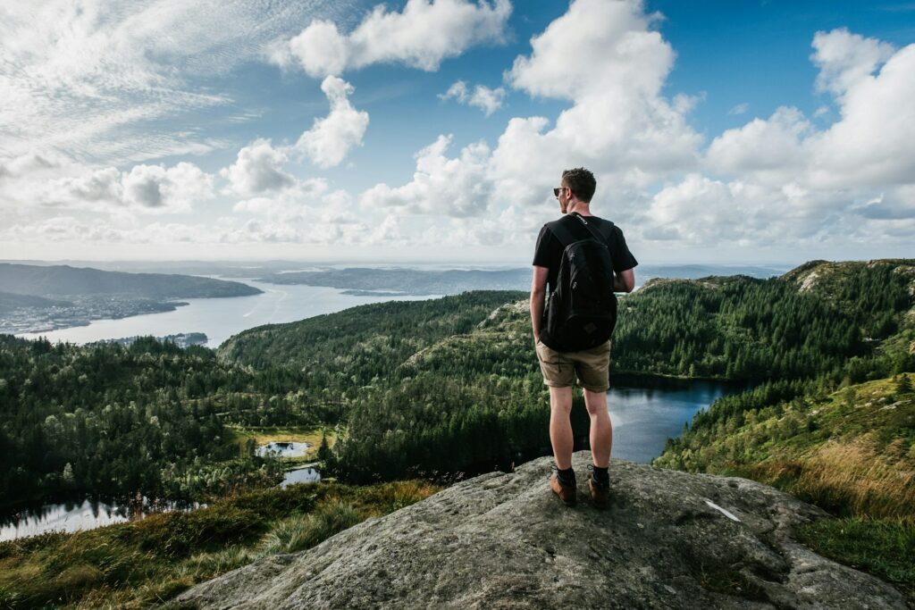 A hiker overlooks a lush, green valley and shimmering lakes, set against a backdrop of clouds and blue skies.
