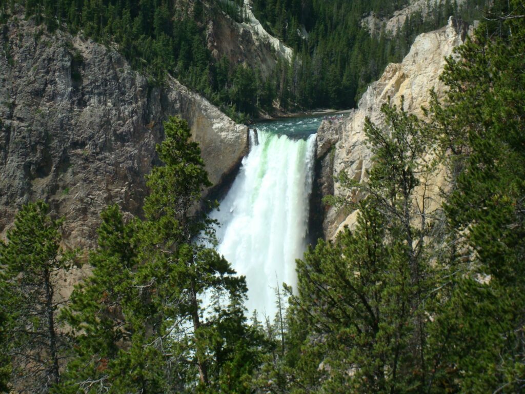 A powerful waterfall cascades down rocky cliffs, surrounded by lush greenery and distant pine trees in a serene natural landscape.