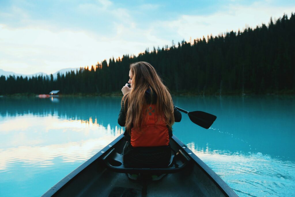 A person with long hair sits in a canoe on a serene turquoise lake, surrounded by lush pine trees and a cloudy sky at dusk.