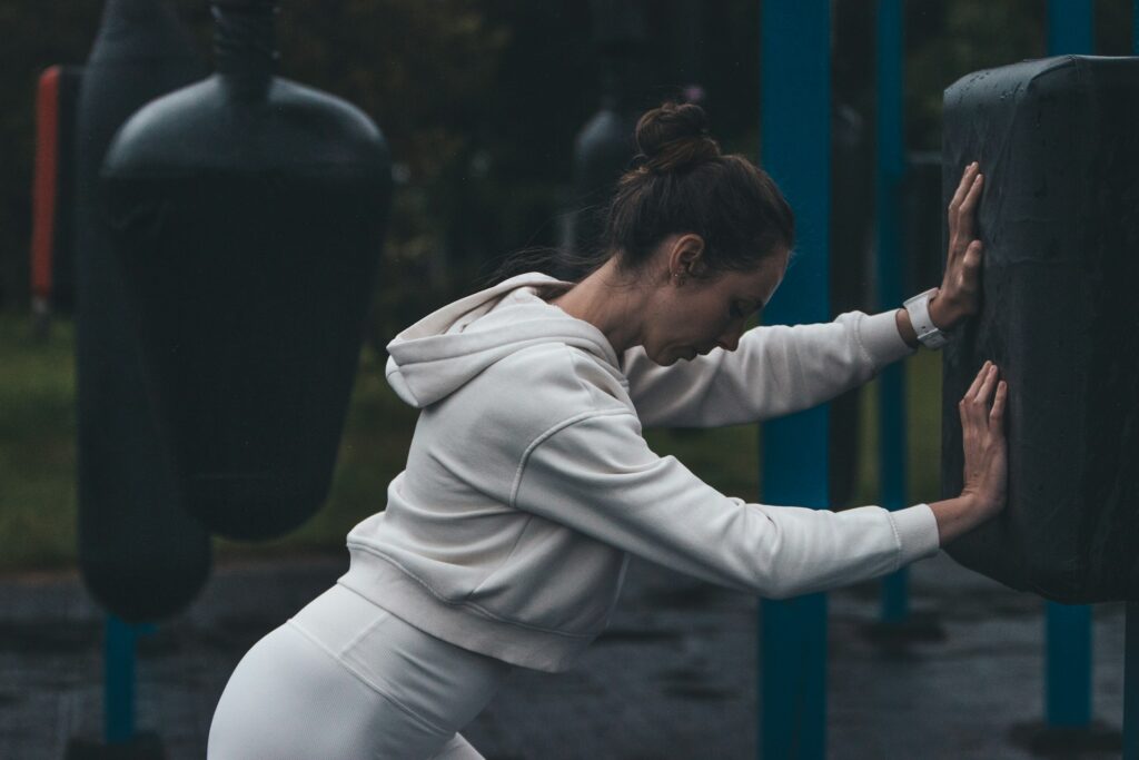 A person in a white hoodie and leggings leans against a heavy bag, engaged in an outdoor workout in a rainy environment.