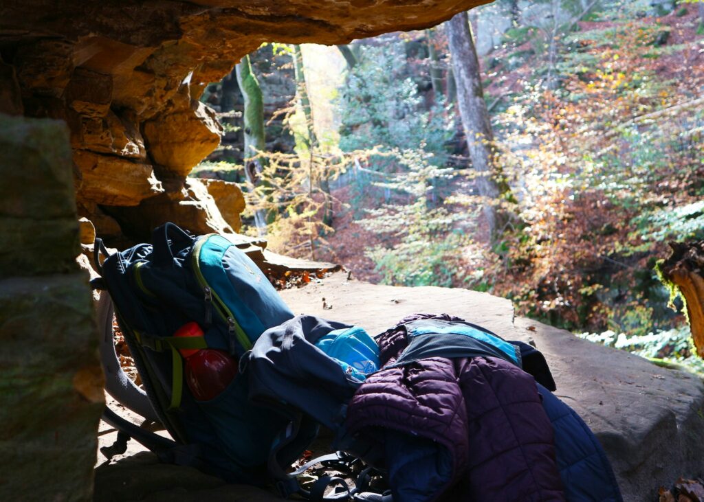 A colorful backpack and folded jackets are nestled in a natural cave, surrounded by a sunlit forest in autumn.