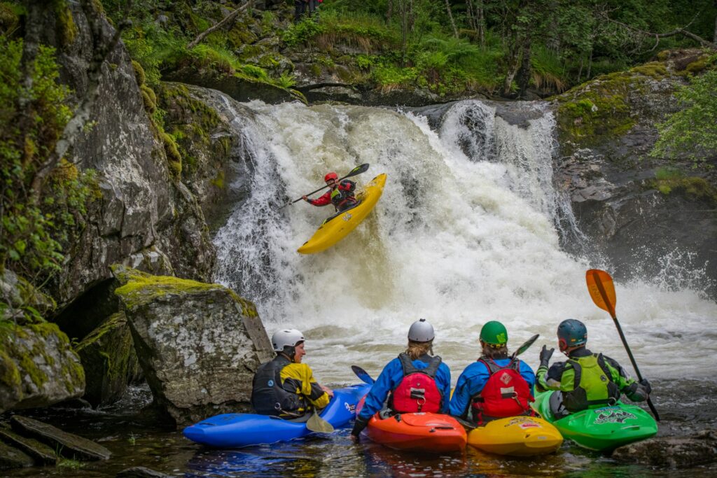 A kayaker navigates a waterfall while three other paddlers observe from the riverbank, surrounded by lush greenery and rocky terrain.