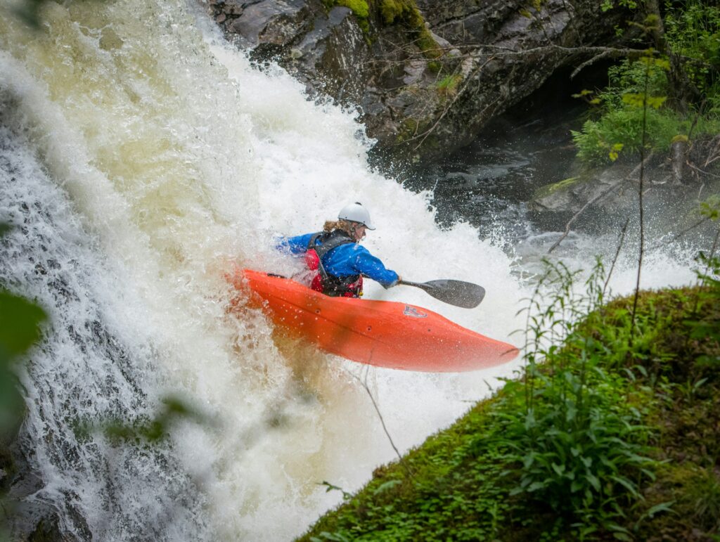 A kayaker in an orange kayak navigates a rushing waterfall, surrounded by lush greenery and splashing water.