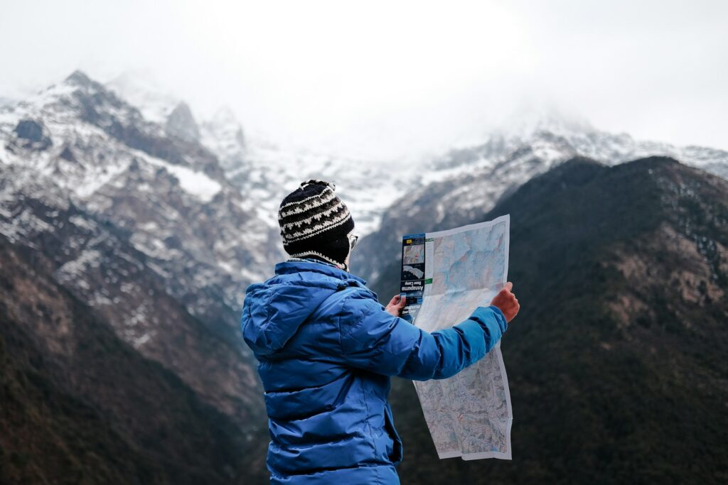 A person in a blue jacket and knit hat studies a map against a backdrop of snow-capped mountains and cloudy skies.