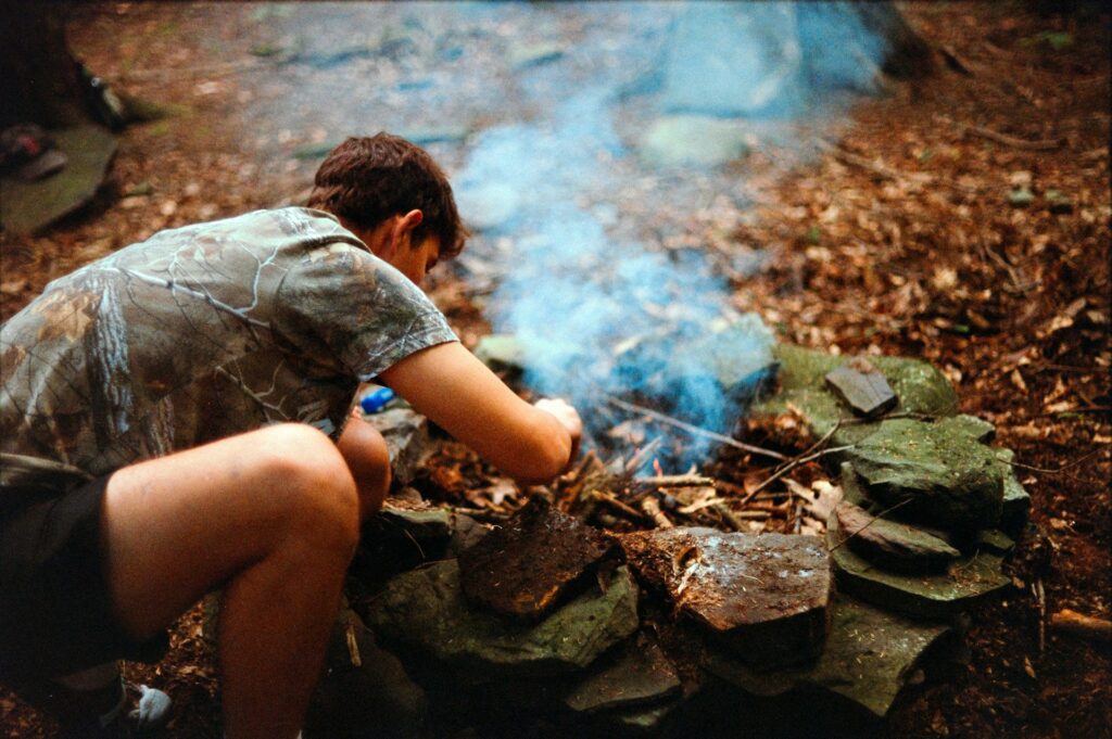 A person in a camouflage shirt crouches by a rustic stone fire pit, tending to a small fire amidst a forested setting.