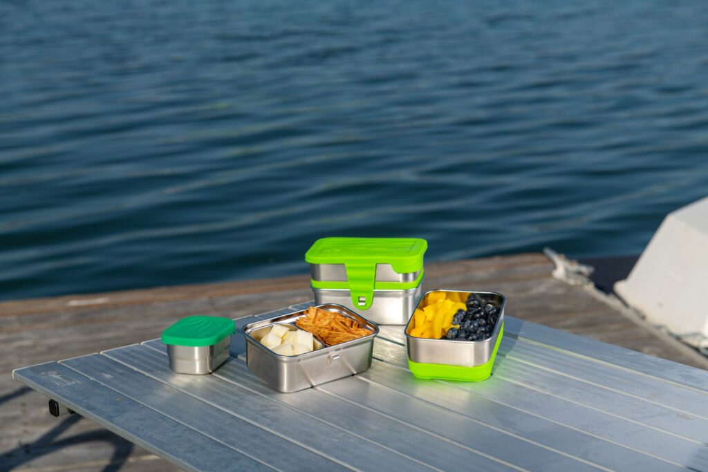 Three stainless steel lunch boxes with green lids, filled with fruit, sit on a table by the water. Bright blue and green color contrast.