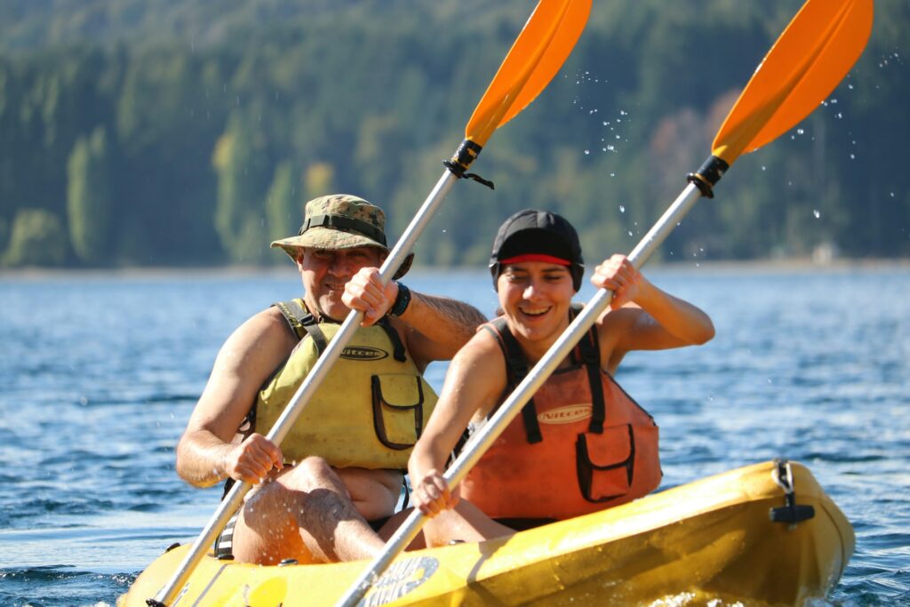 A couple kayaks together on a sunny day, wearing life jackets and paddling on a tranquil lake surrounded by green trees.