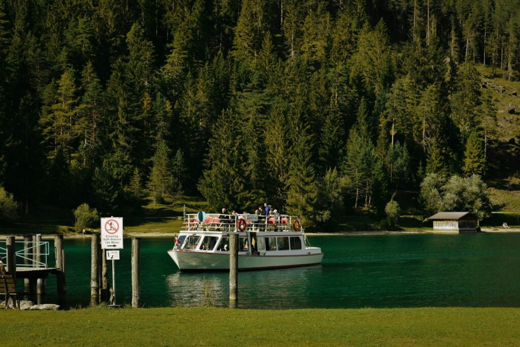 A boat with passengers glides across a clear lake, surrounded by lush green trees and a rustic wooden shed in the background.