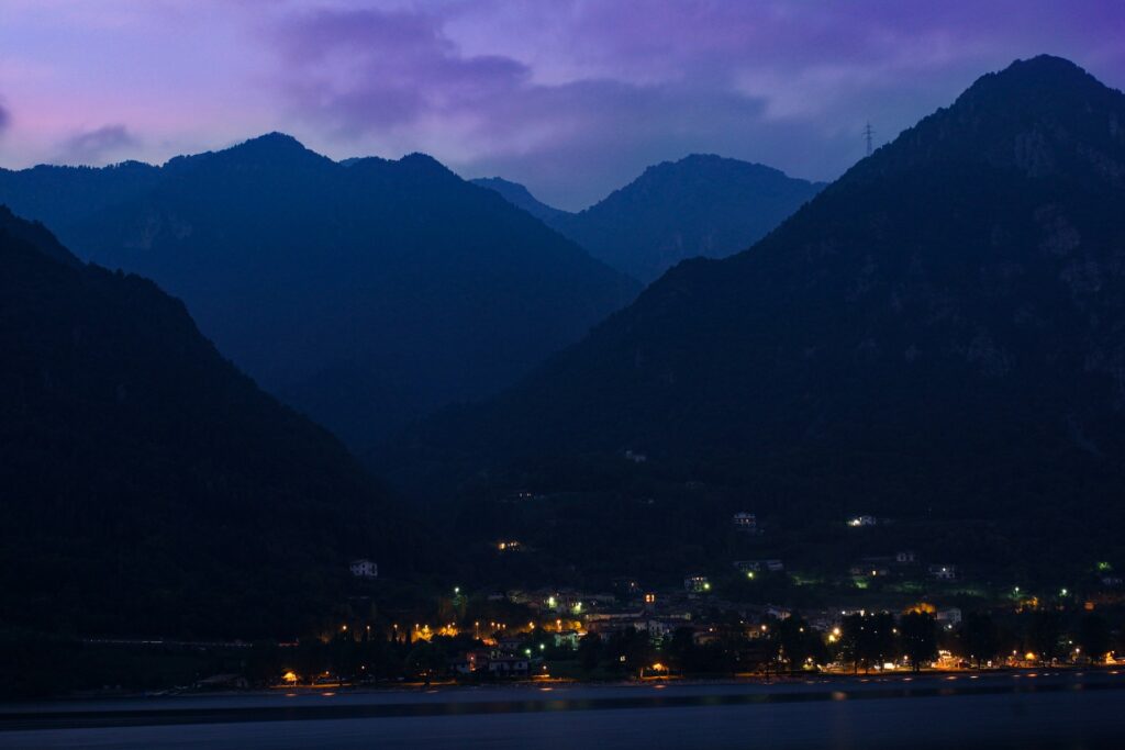 Evening view of a small village nestled among dark mountains, illuminated by warm lights under a purple-hued sky.