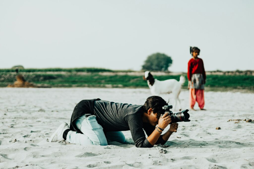 A man photographing a goat on the beach, with waves and sand in the background.