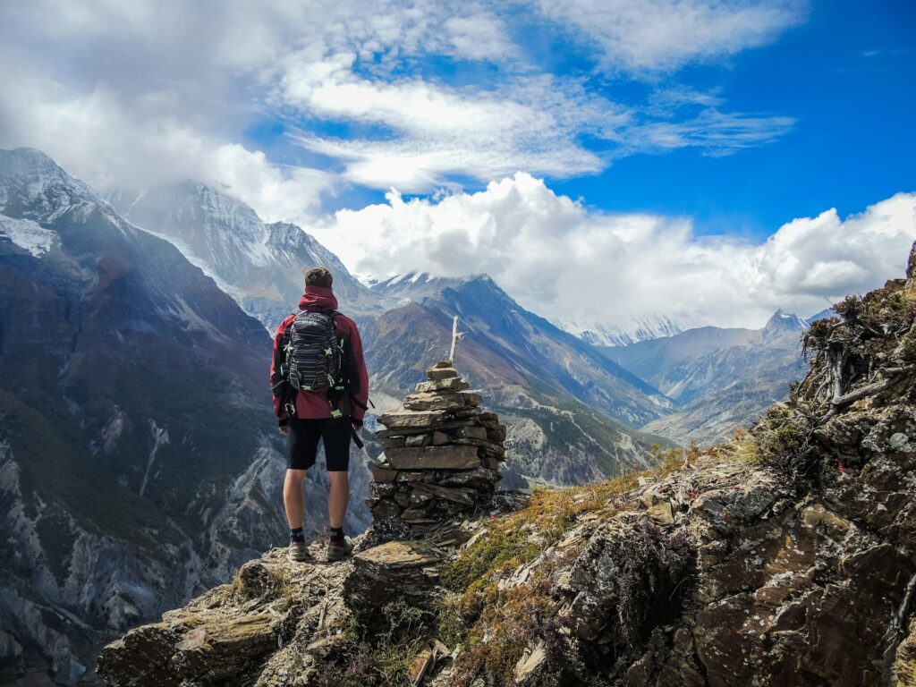 A hiker stands atop a mountain ledge, gazing at a stunning valley and snow-capped peaks beneath a partly cloudy sky.