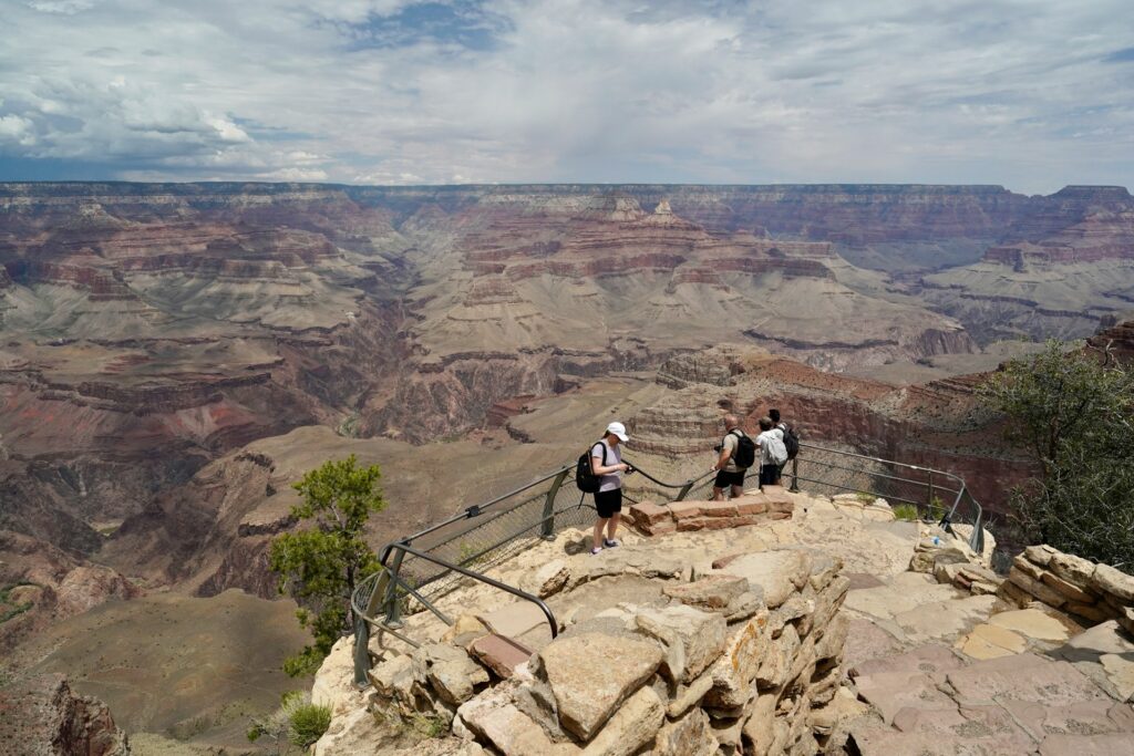 Three hikers stand on a viewing platform at the Grand Canyon, admiring the expansive, layered landscape under a partly cloudy sky.