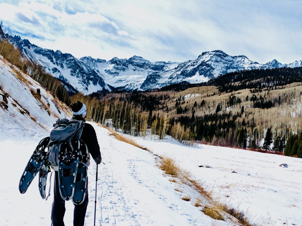 A hiker with a backpack and snowshoes walks along a snowy trail, surrounded by towering mountains and a forested valley.