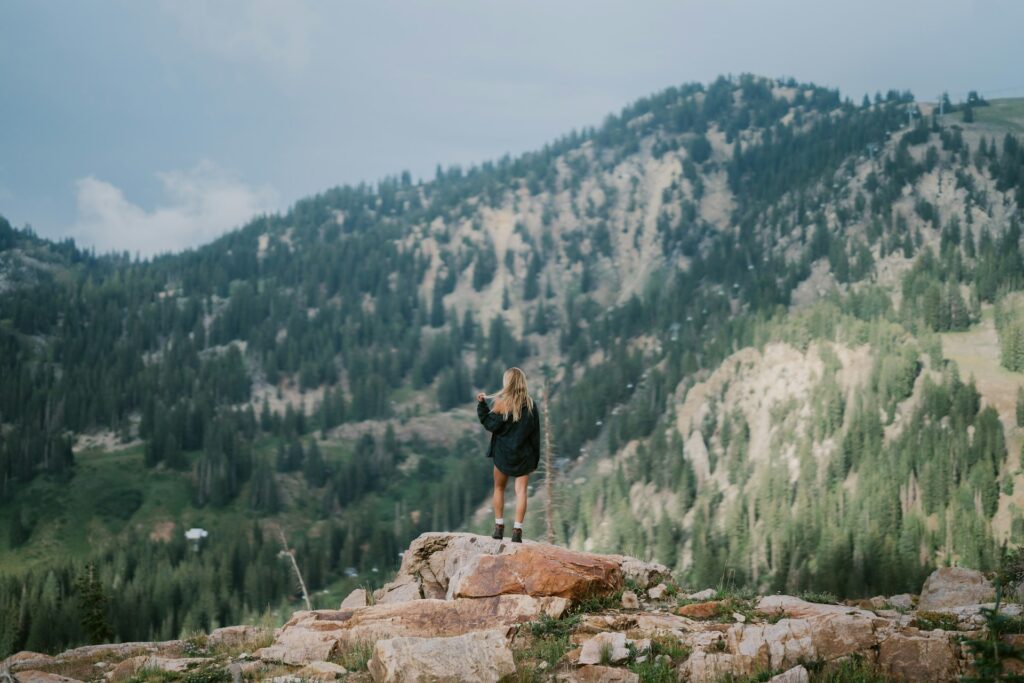 A woman with long hair stands on a rock, gazing at lush green mountains under a cloudy sky.