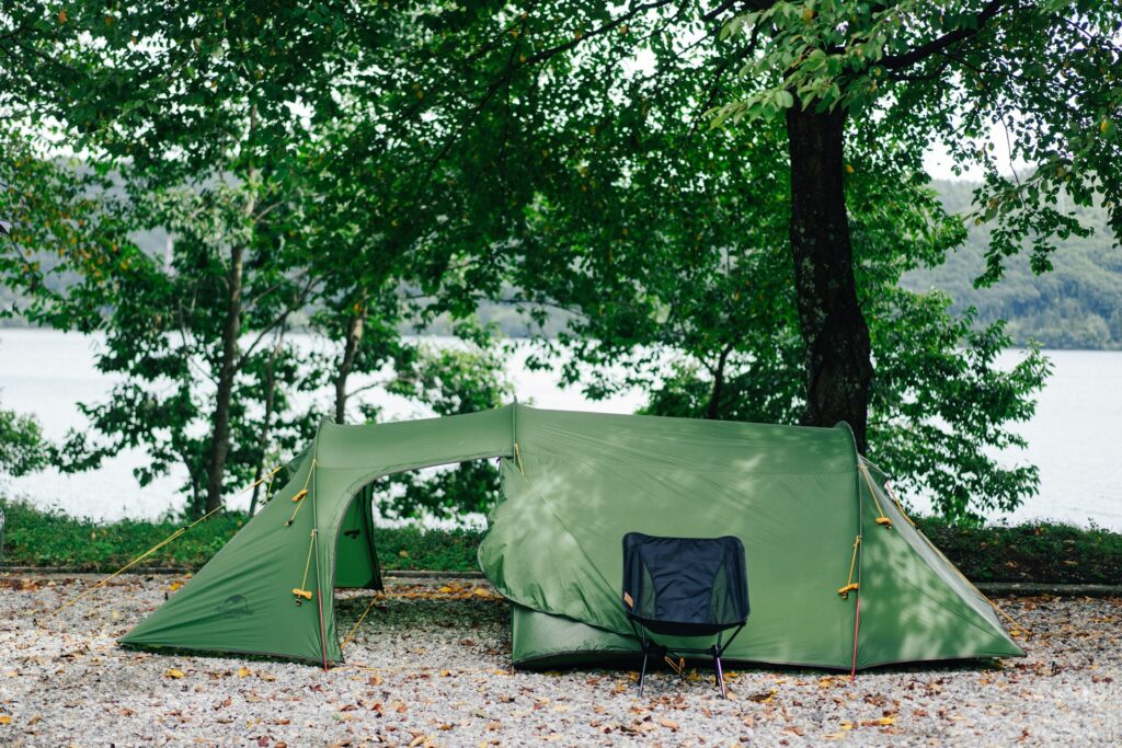 A green camping tent with an open entrance sits on gravel under trees, next to a black camping chair by a lake.