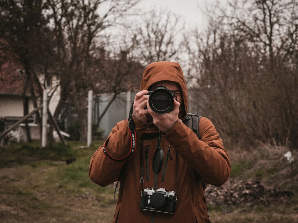 A person in an orange hooded jacket stands outdoors, holding a camera, surrounded by trees and a grassy area.