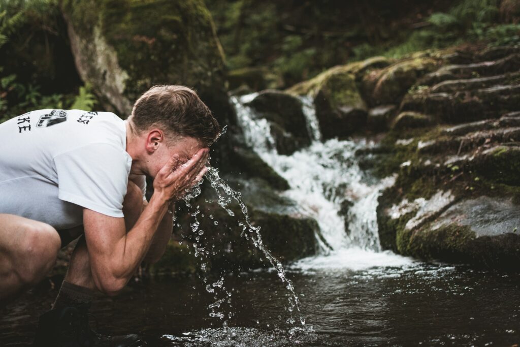 A man crouches by a tranquil stream, splashing water on his face amidst lush forest greenery.