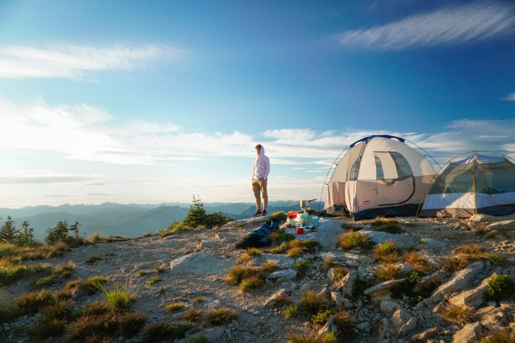 A person stands on a rocky mountain overlook by two tents, surrounded by nature and breathtaking views of distant peaks.
