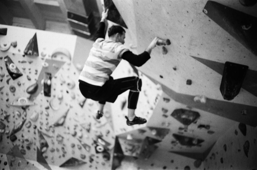 A climber in a striped shirt ascends a challenging indoor rock wall, surrounded by various climbing holds. Black and white photograph.