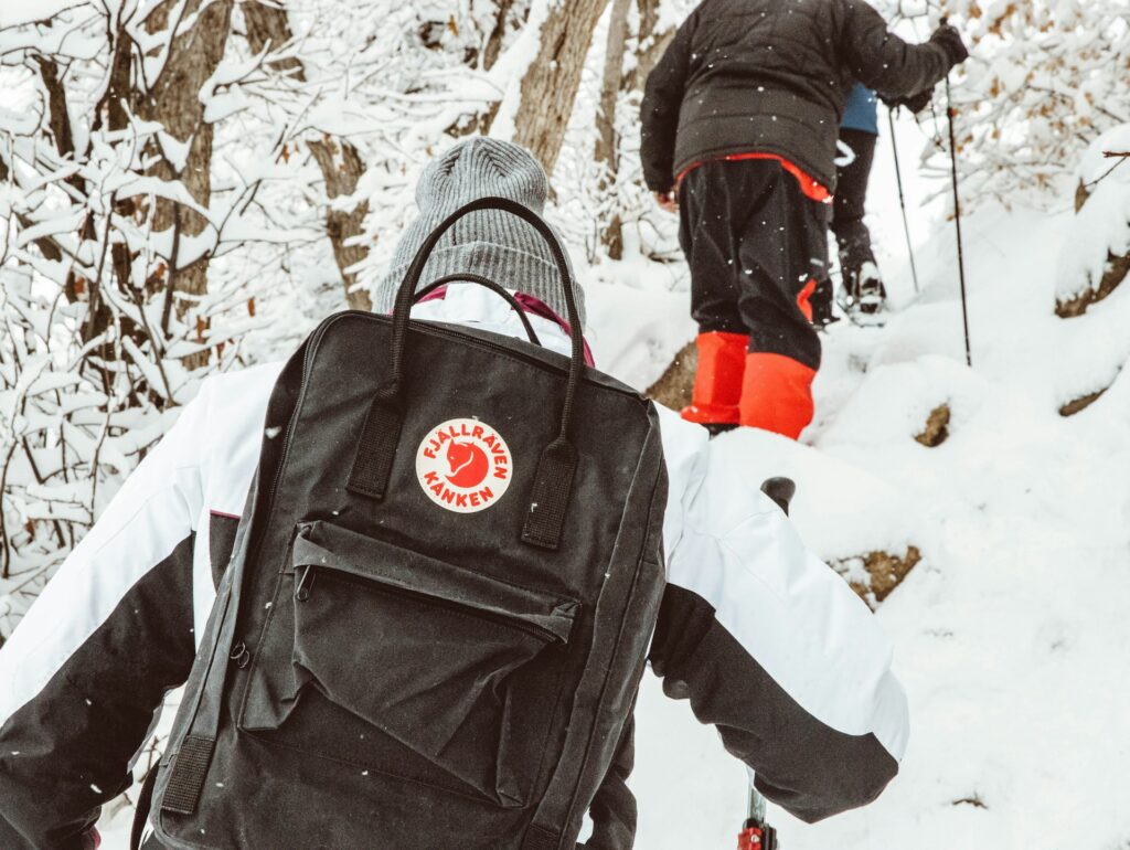 A person with a black Fjällräven Kanken backpack climbs a snowy trail, while another hiker in red pants follows behind.