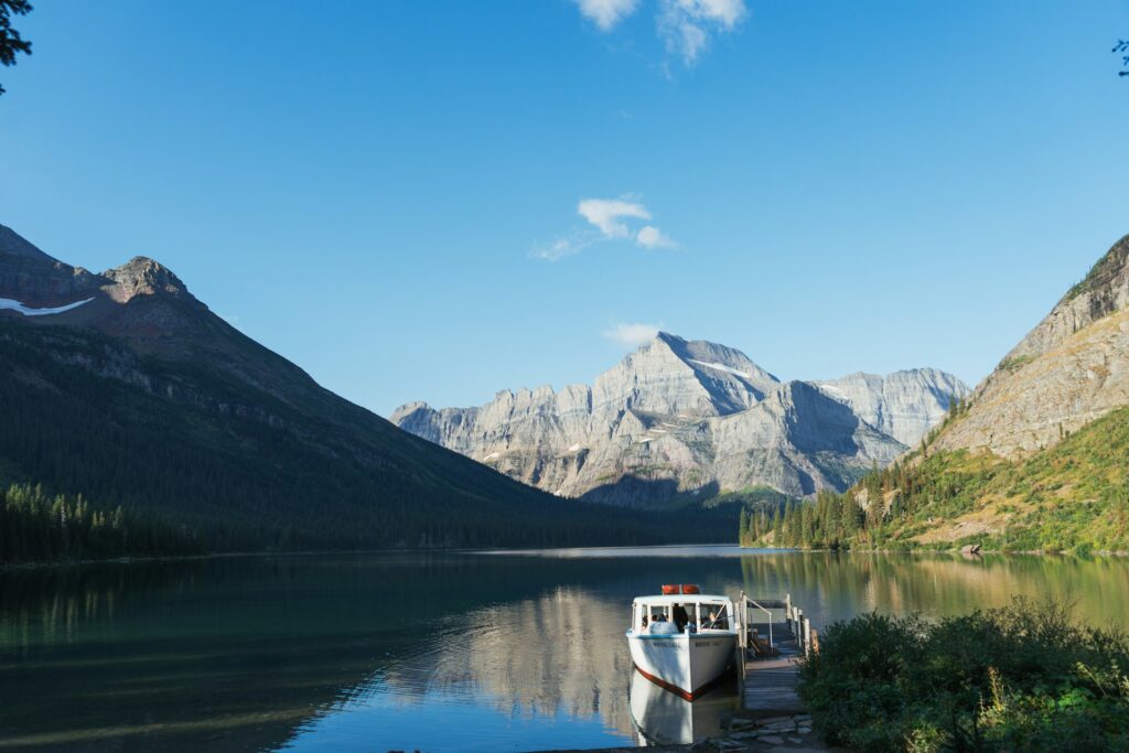 A tranquil lake scene with a boat docked at a wooden pier, surrounded by towering mountains and lush forest under a clear blue sky.