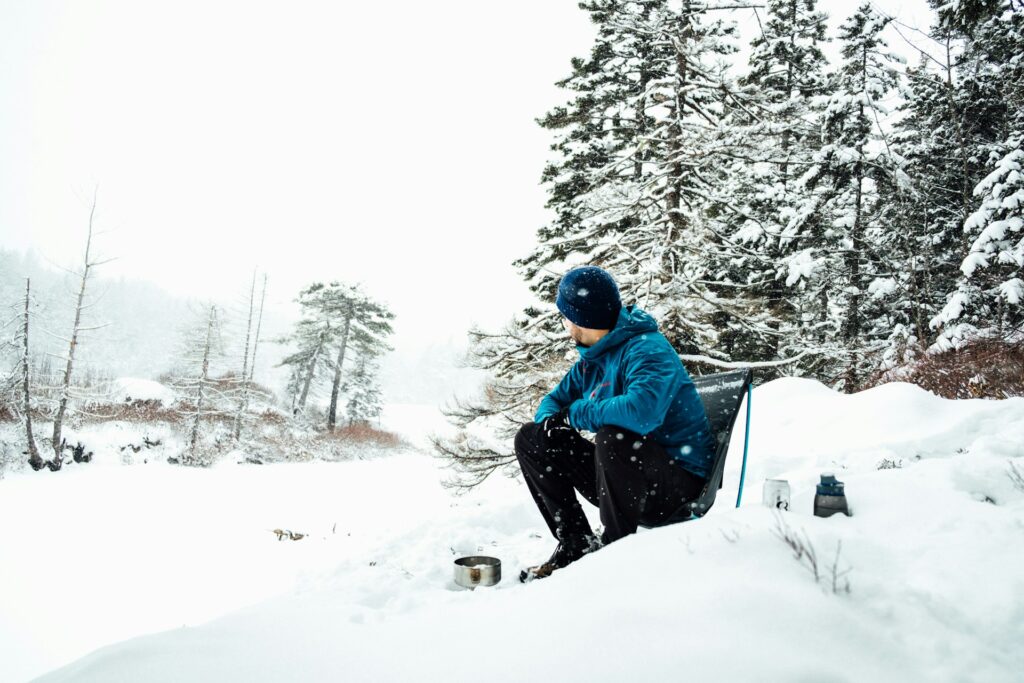 A man in a blue jacket and beanie sits on a snowy bank, gazing contemplatively at a cooking pot amidst a winter wonderland.