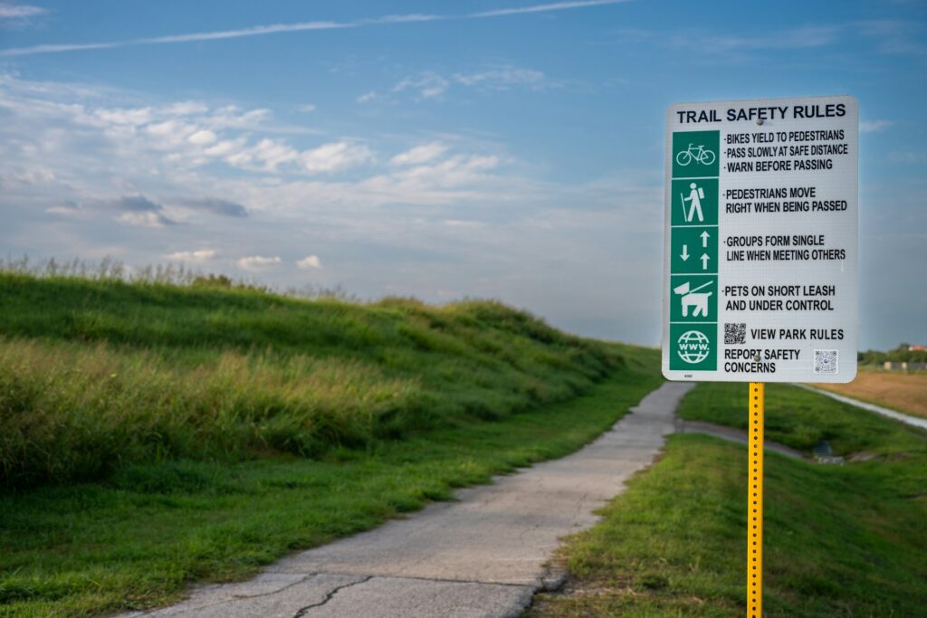 A trail safety rules sign stands beside a paved path surrounded by grassy hills under a blue sky with scattered clouds.