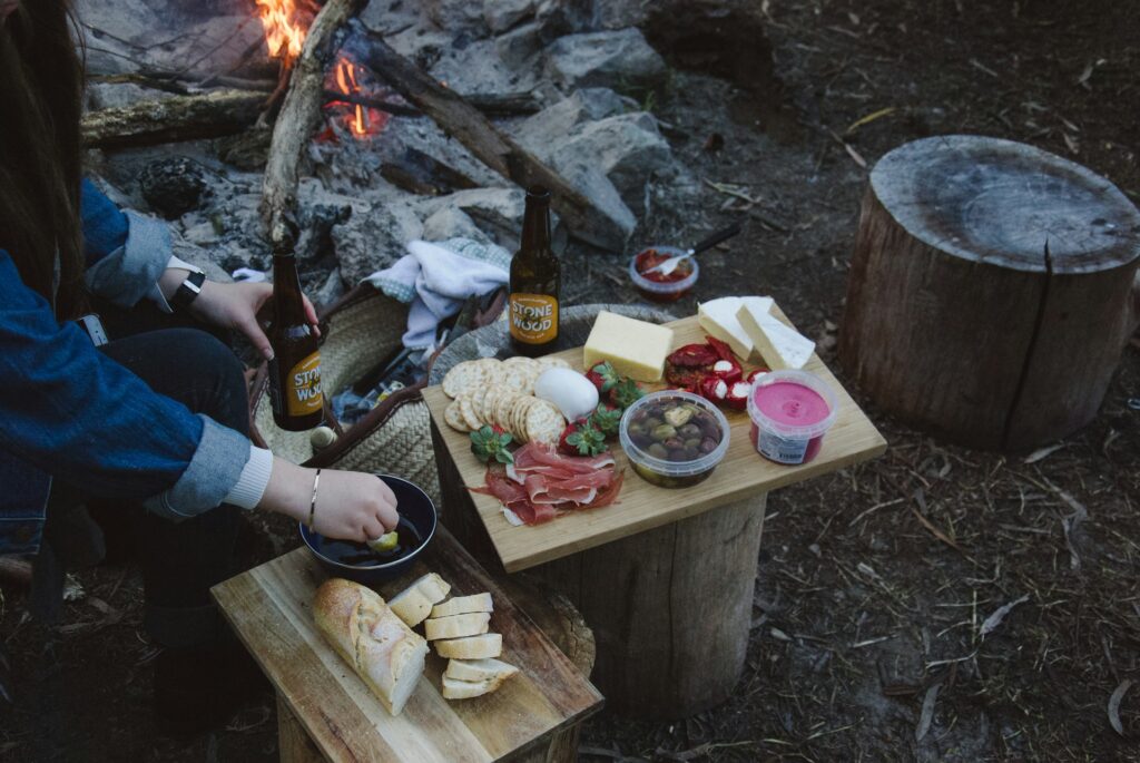 A cozy outdoor picnic features cheese, charcuterie, bread, and drinks near a campfire, with a person enjoying a snack.