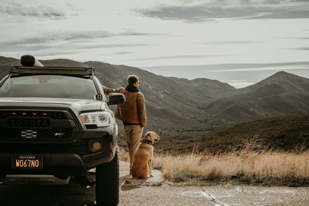 A man and his golden retriever stand beside a Toyota truck, gazing at a scenic mountain landscape under a cloudy sky.