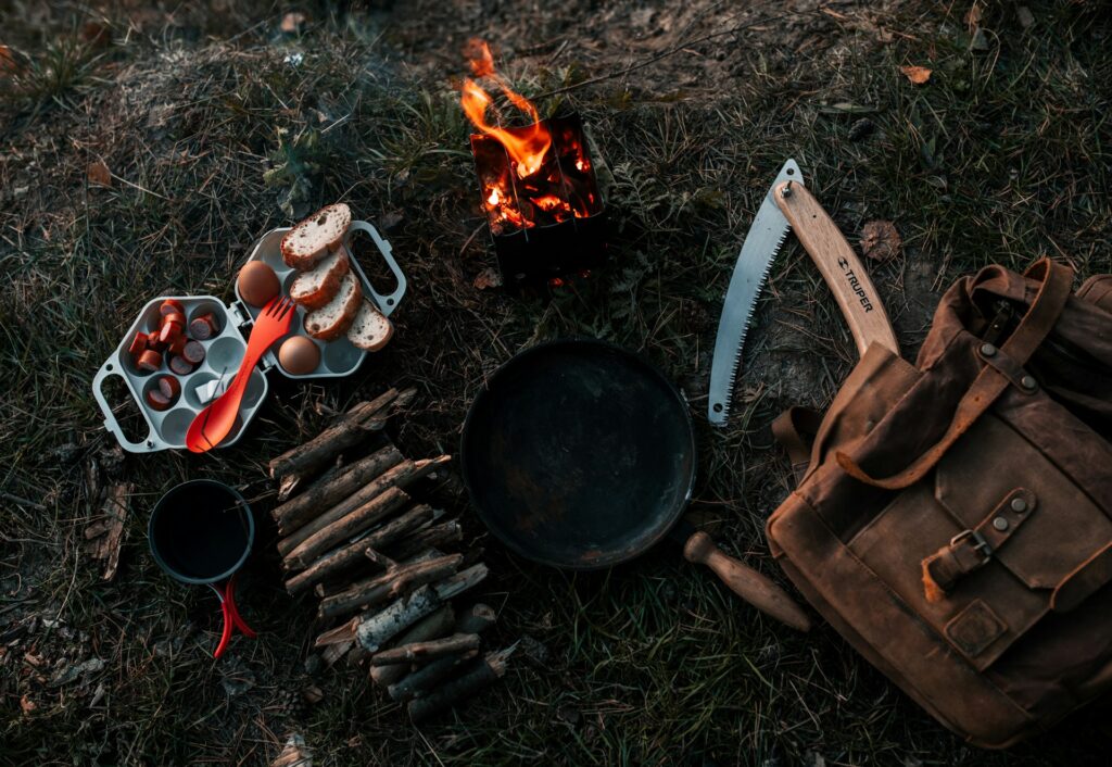 A camping setup featuring a small fire, cooking utensils, food items, and a saw atop grass, with a canvas backpack nearby.
