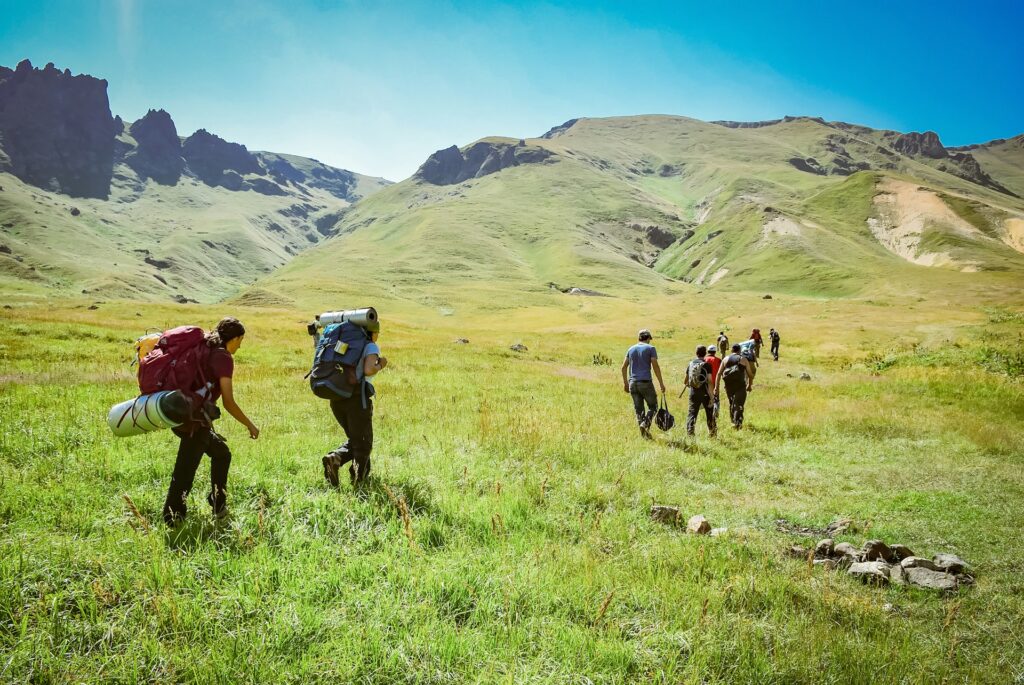 A group of hikers with backpacks trekkers through lush green meadows, surrounded by towering mountains under a clear blue sky.