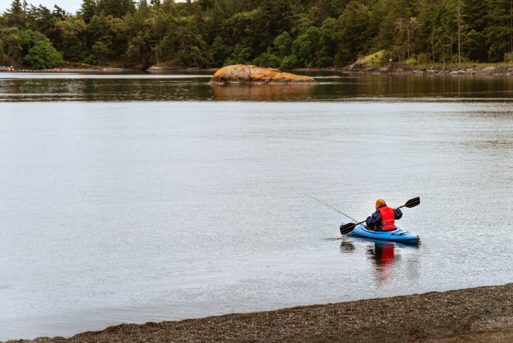 A person in a blue kayak, wearing a red life jacket and hat, fishes calmly on a serene lake, surrounded by green trees and distant rocks.
