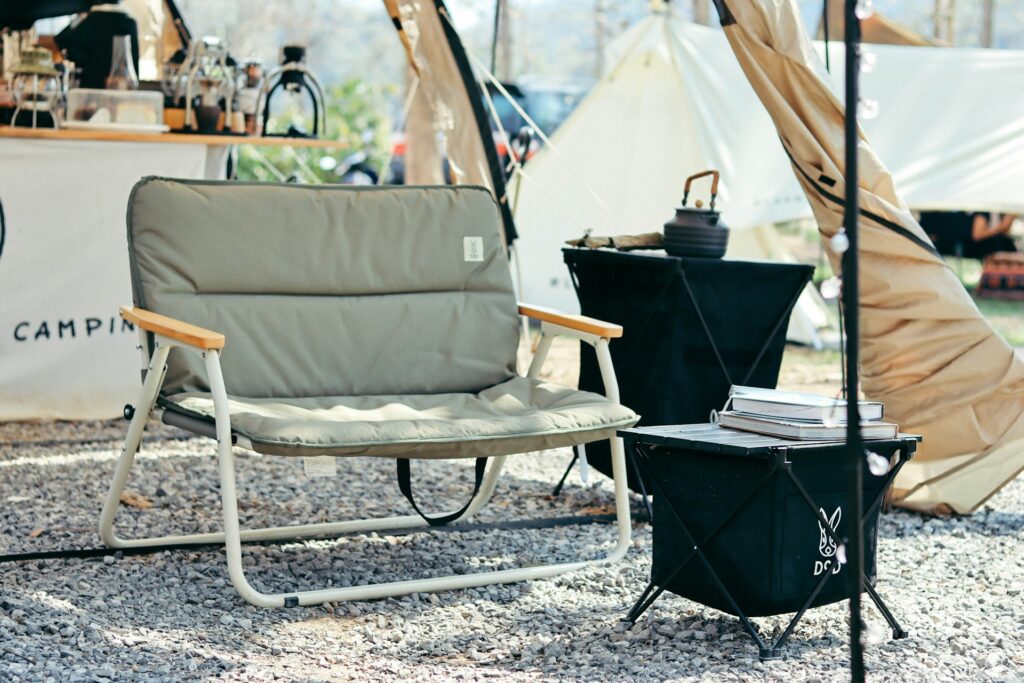 A cozy camping setup featuring a lightweight, gray folding chair with wooden armrests next to a small black side table stacked with books.