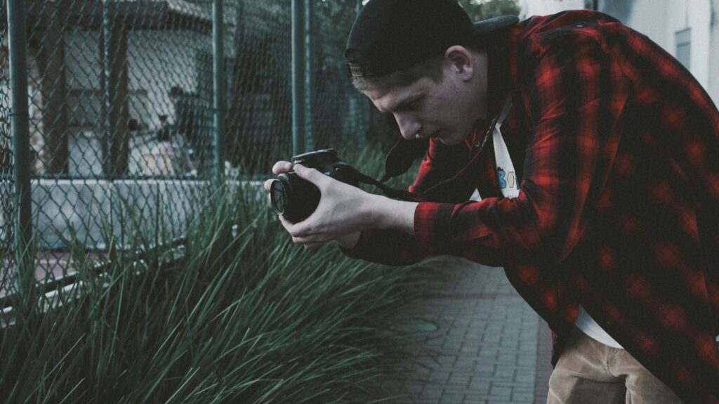 A person in a red and black flannel shirt kneels, focusing a camera on lush green grass near a fenced area.