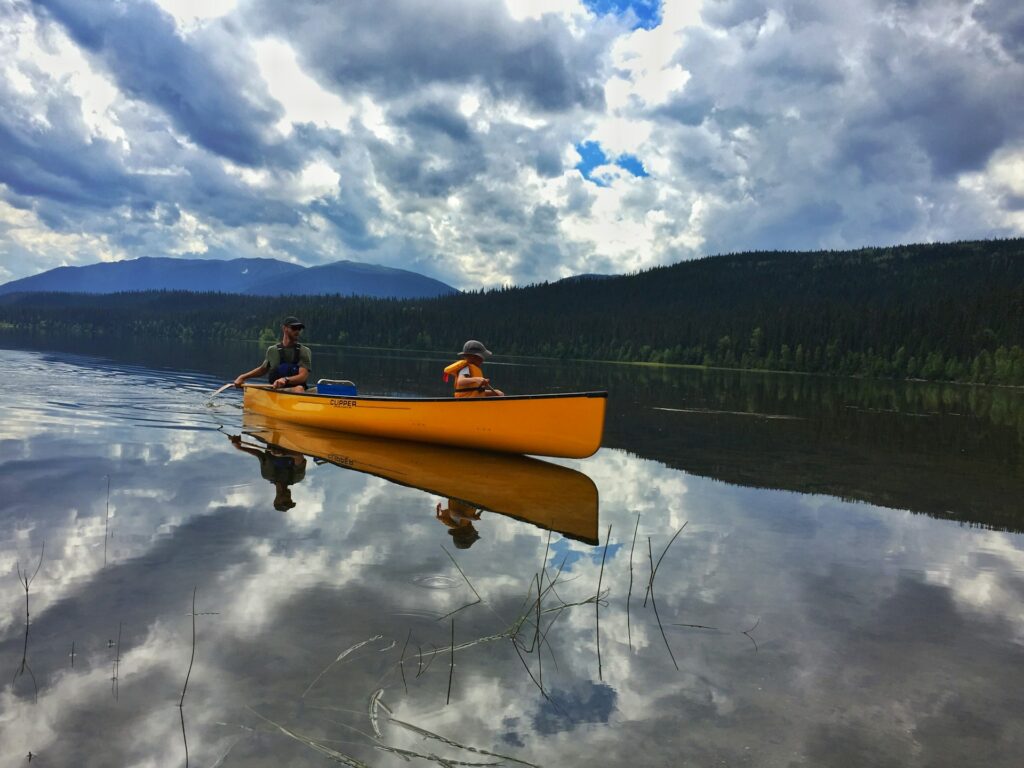 Two people paddle a bright yellow canoe on a calm lake, surrounded by lush greenery and dramatic cloud reflections.