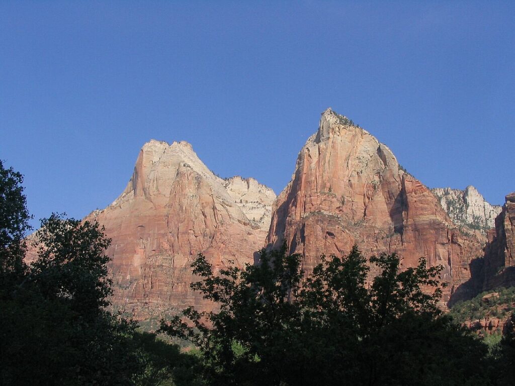 Majestic red rock formations rise against a clear blue sky, framed by lush green trees in Zion National Park.