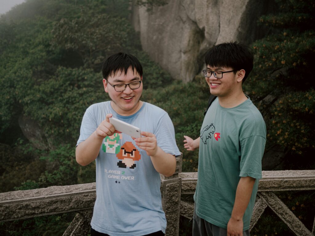 Two young men stand on a rocky cliff surrounded by lush greenery, one holding a smartphone while the other gestures animatedly.