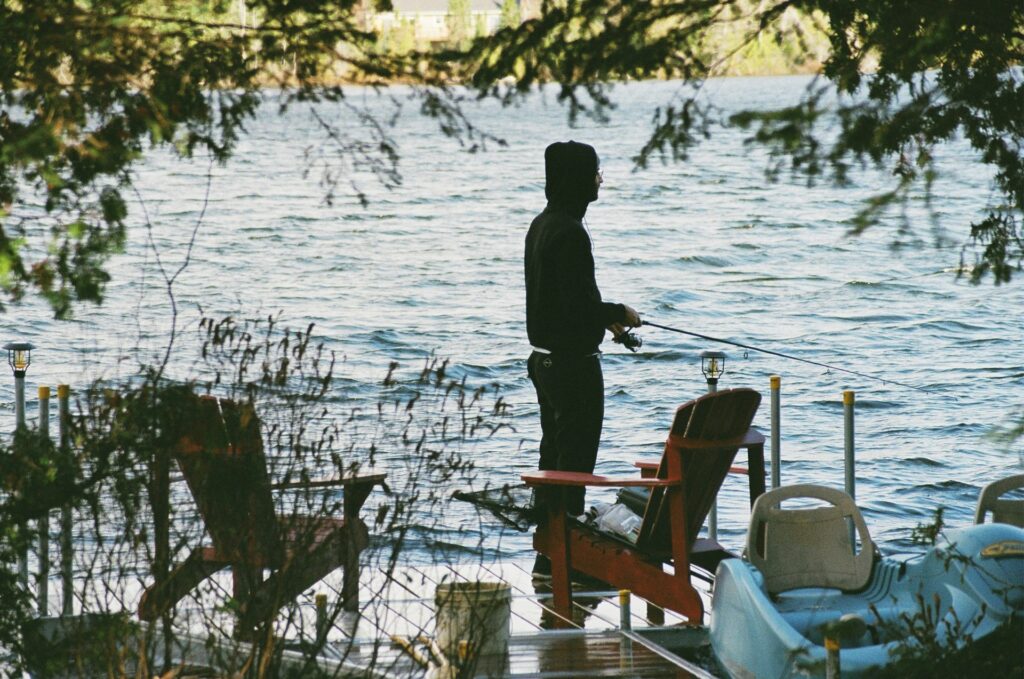 A person in a black hoodie fishes by the water's edge, surrounded by trees and a bright red chair on a serene dock.