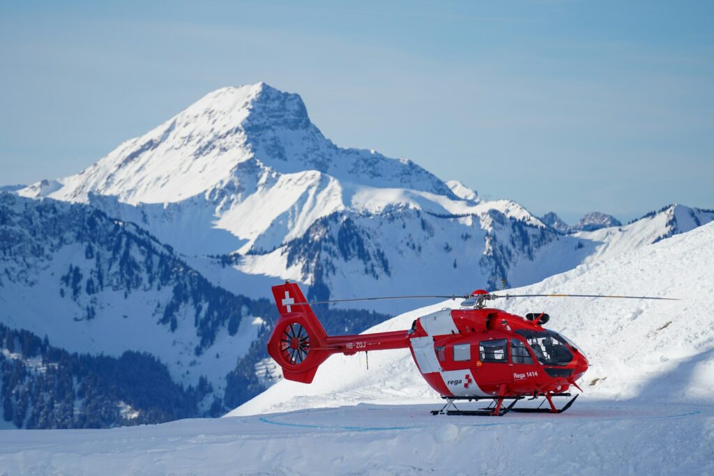 A red Swiss helicopter parked on snow with majestic mountains in the background under a clear blue sky.