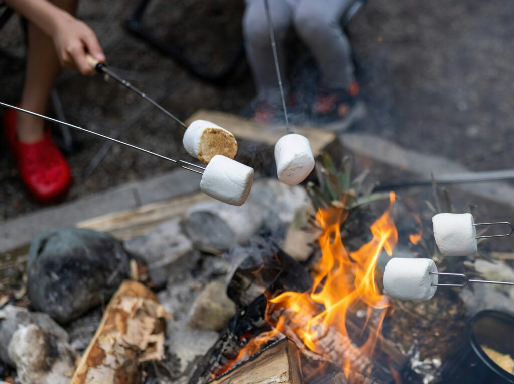 Close-up of marshmallows roasting over a campfire on skewers, with a blurred figure in the background, and glowing flames.
