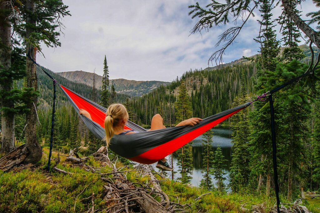 A woman relaxes in a red and gray hammock nestled among tall trees, overlooking a serene lake and mountain landscape.