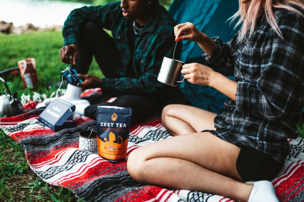 A picnic scene featuring two people by a tent, enjoying Zest Tea on a colorful blanket by the water's edge.