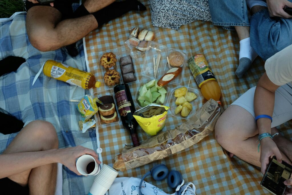 A colorful picnic spread on a checkered blanket, featuring snacks, drinks, and fresh fruit, with friends enjoying the meal around it.