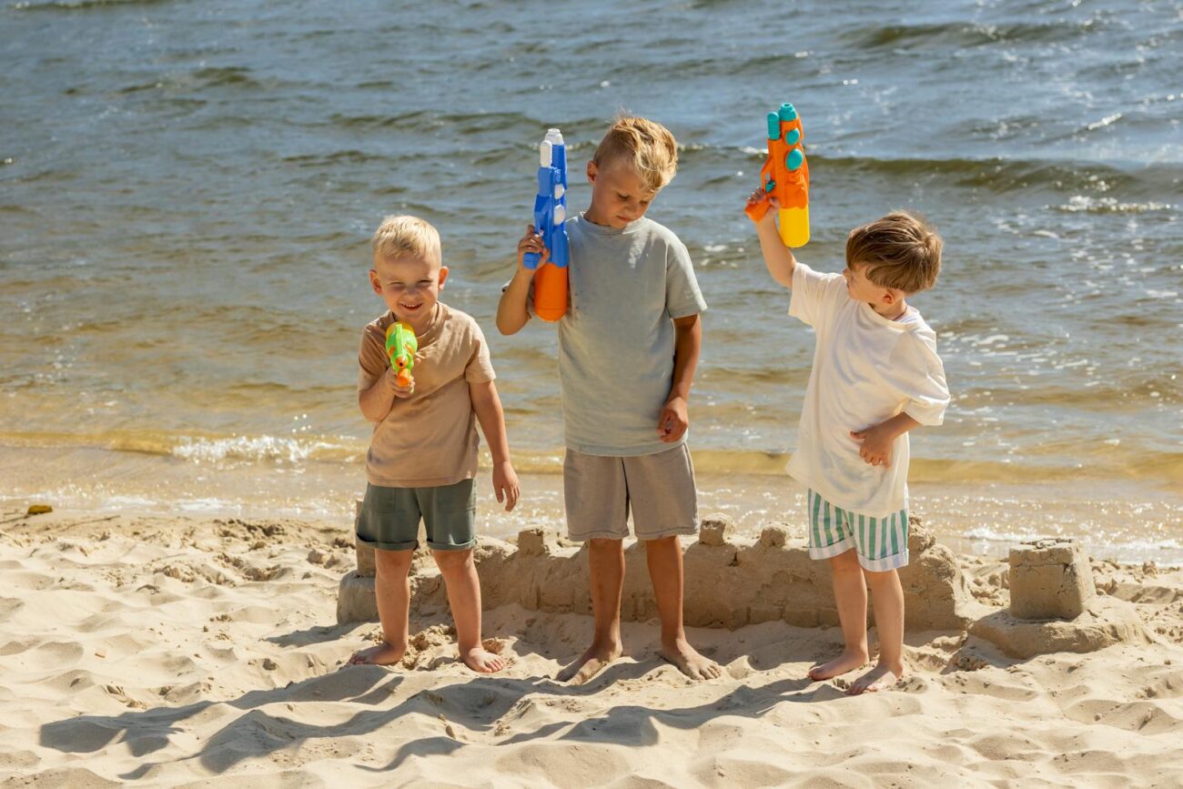 Three boys play with water guns on a sandy beach during a sunny summer day.