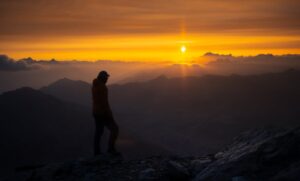 A solitary hiker silhouetted against a vibrant sunset in a mountainous landscape.