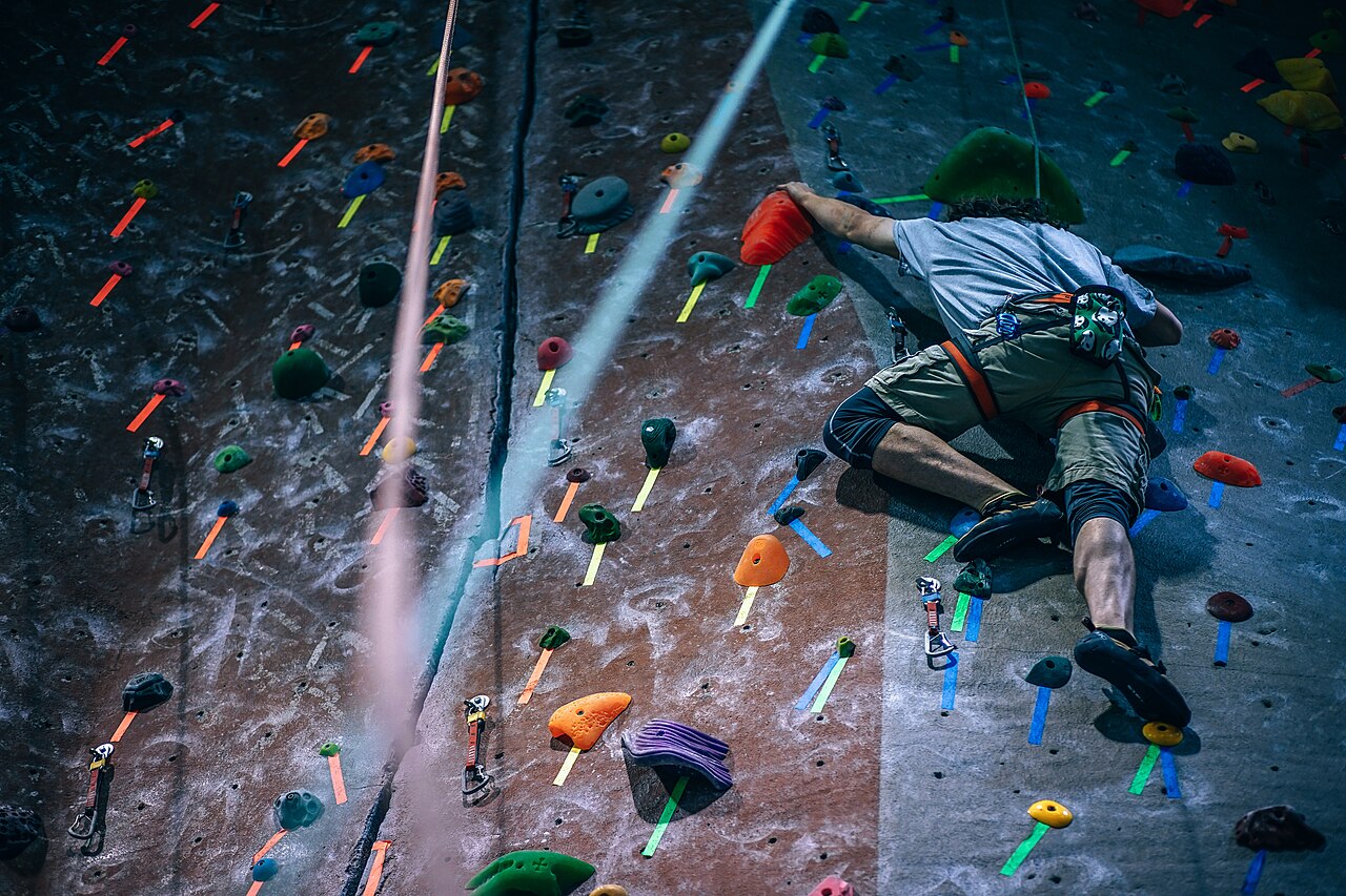 A climber ascends a colorful indoor rock wall, using vibrant holds and secured by a rope for safety.